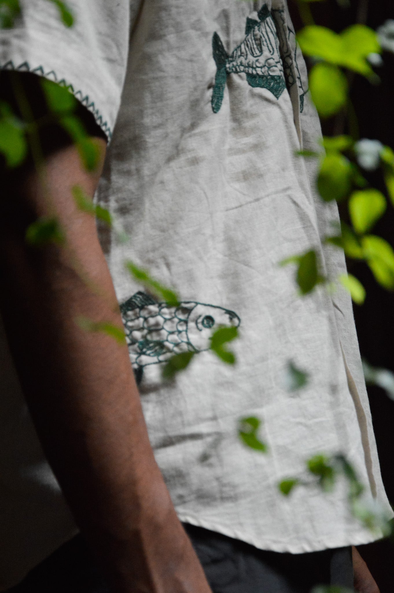 details of a half-sleeved beige linen shirt with a detailed green fish embroidered across the centre of the shirt  and a smaller fish peeking from the left bottom corner