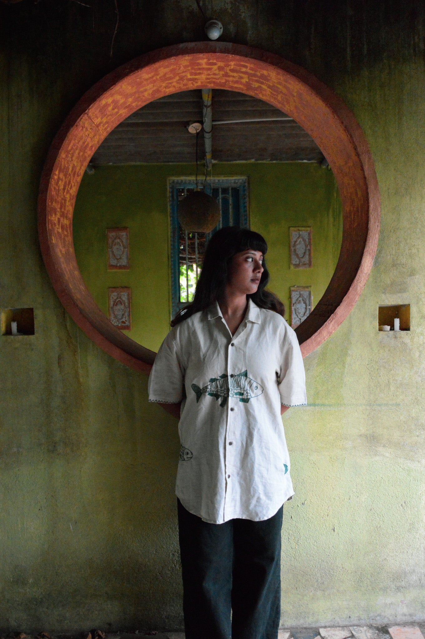 A girl in half-sleeved beige linen shirt with a detailed green fish embroidered across the centre of the shirt is standing against a mustard yellow wall