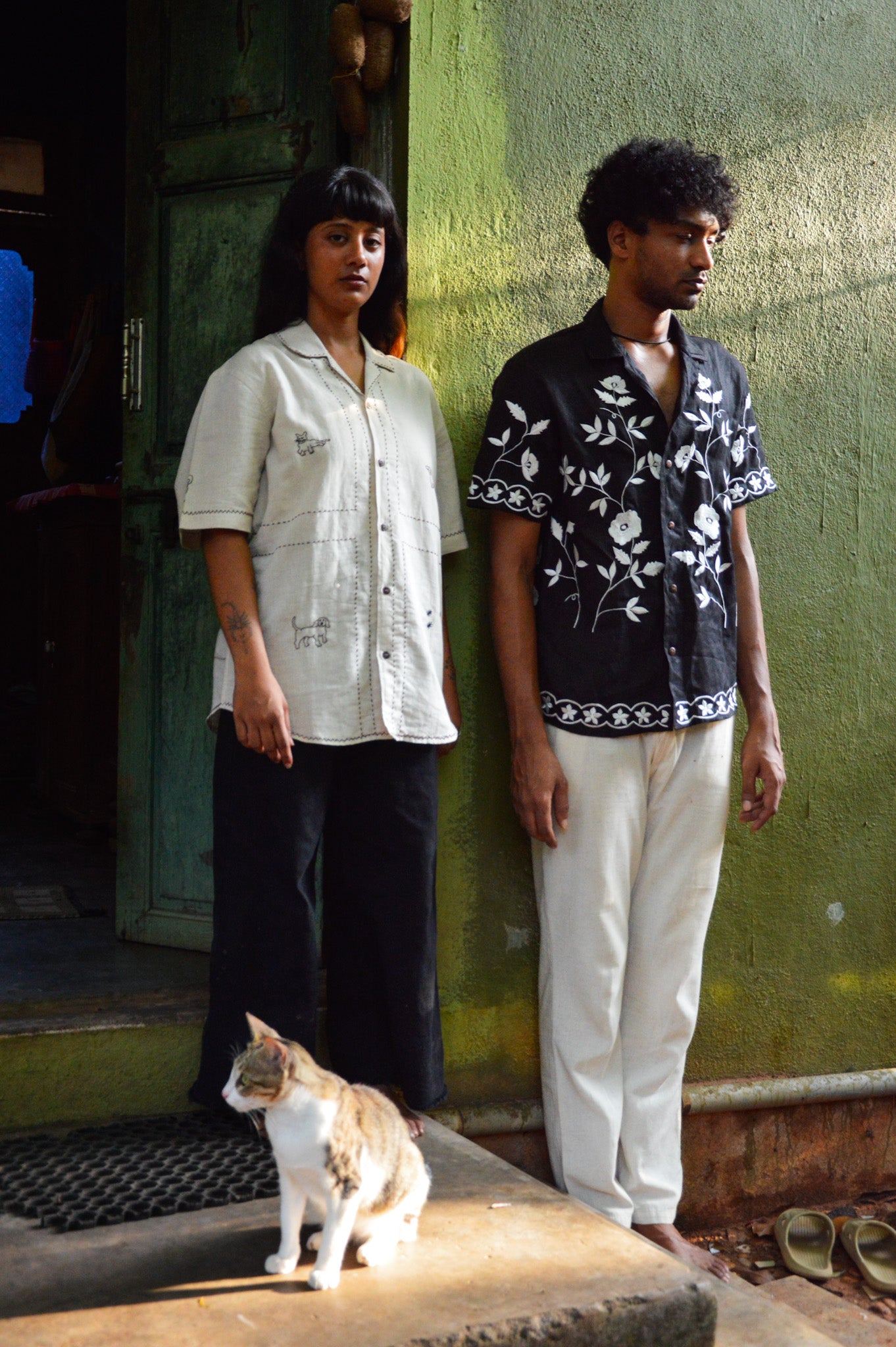 A young man in a black linen half sleeved shirt with off-white floral embroidery throughout is standing against a green wall