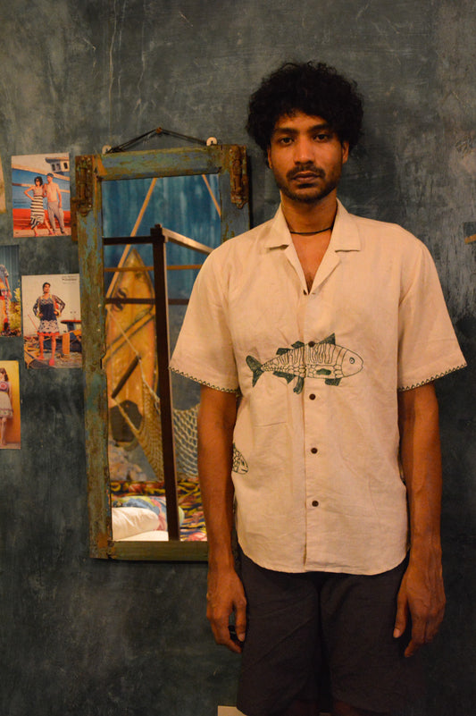 A young man in half-sleeved beige linen shirt with a detailed green fish embroidered across the centre of the shirt is standing against a blue wall