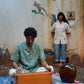 A young man in green linen shirt with flowers embroidered throughout is playing the harmonium