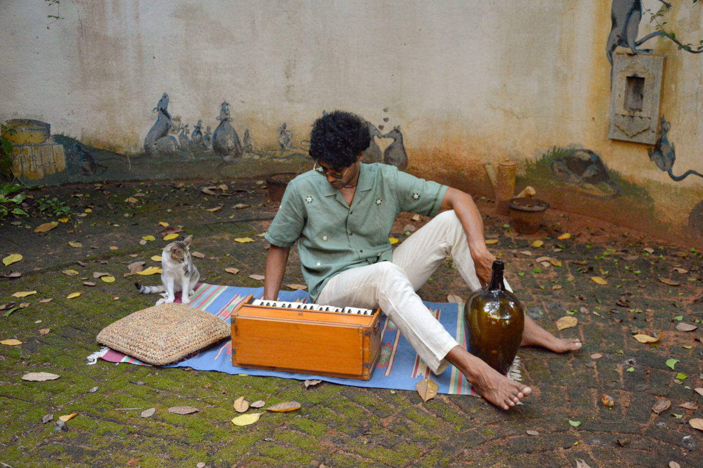 A young man in green linen shirt with flowers embroidered throughout is sitting next to a harmonium