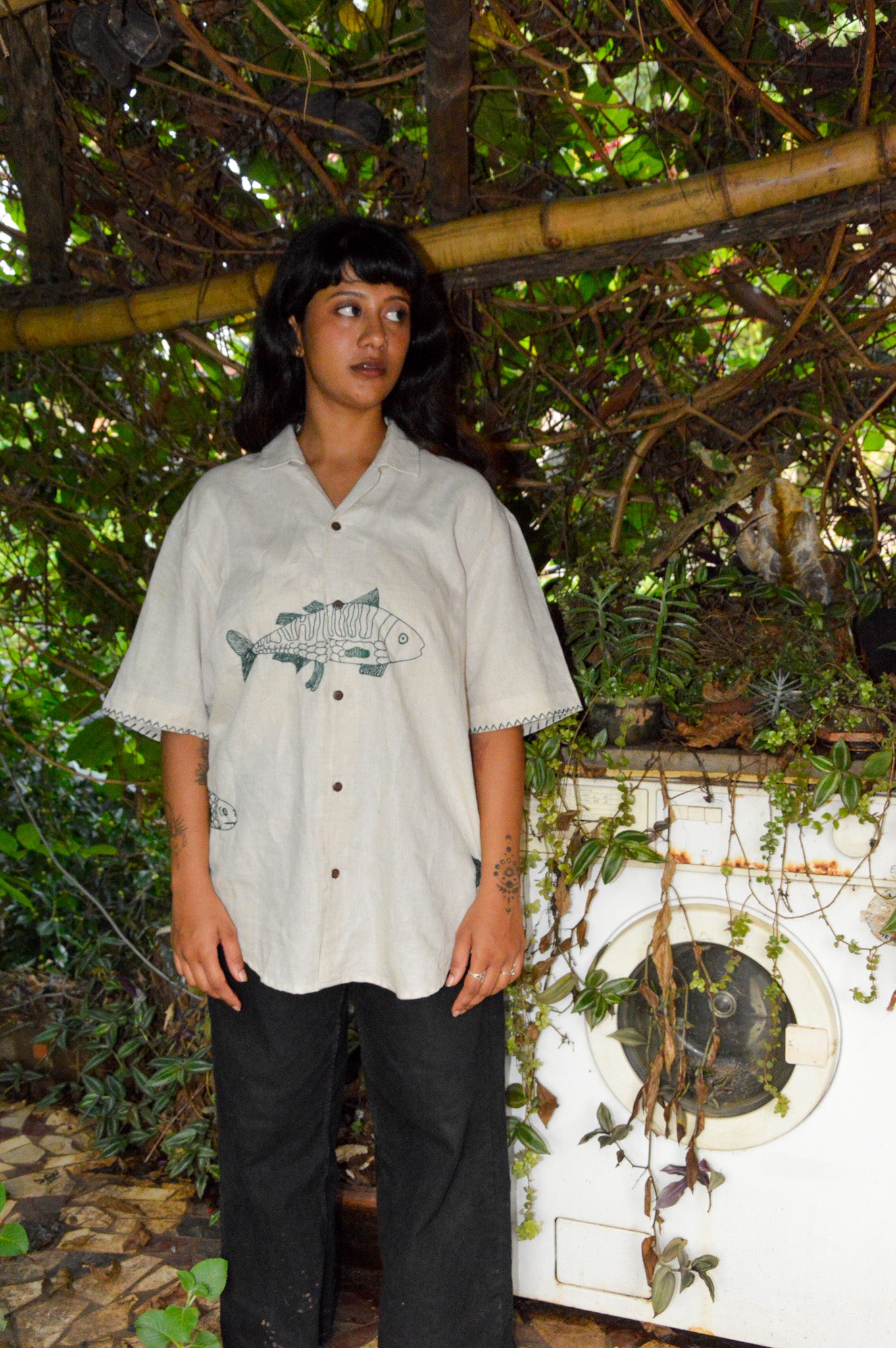 A girl in half-sleeved beige linen shirt with a detailed green fish embroidered across the centre of the shirt is standing in a outdoor kitchen space