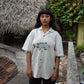 A girl in half-sleeved beige linen shirt with a detailed green fish embroidered across the centre of the shirt is standing outside a hay thatched hut