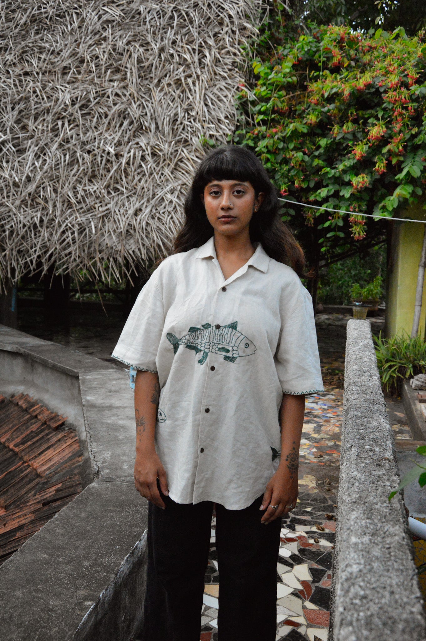 A girl in half-sleeved beige linen shirt with a detailed green fish embroidered across the centre of the shirt is standing outside a hay thatched hut