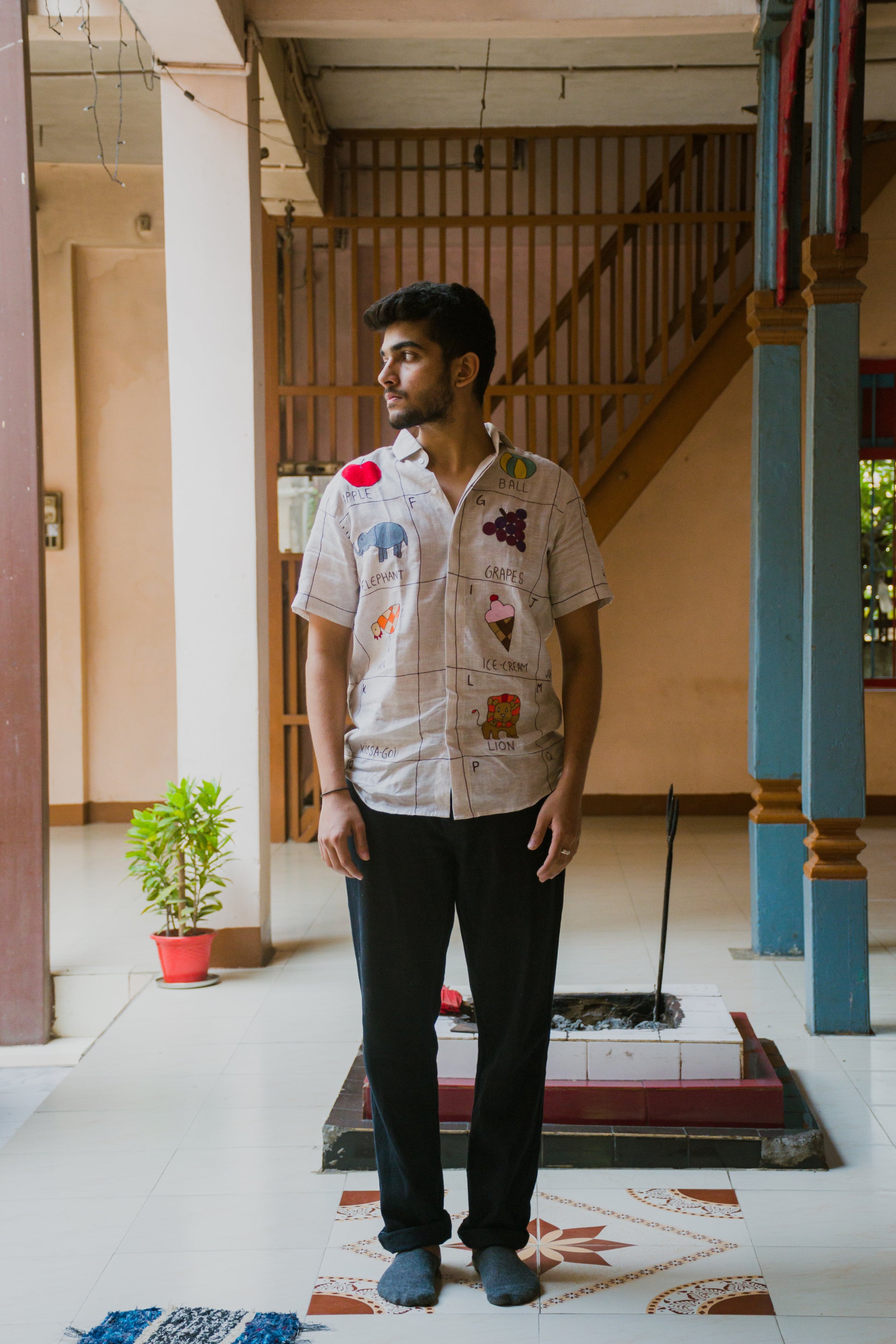 A young man in an beige linen shirt with the hand-embroidered alphabet is standing against a backdrop of a stairs