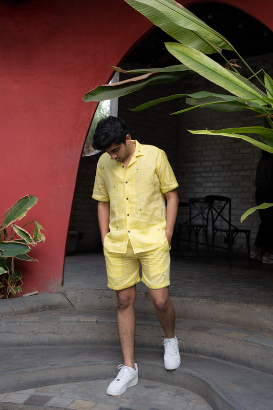 a front view of a man wearing co-ordinated pair of yellow linen shirt and shorts with green sun embroidery at a cafe