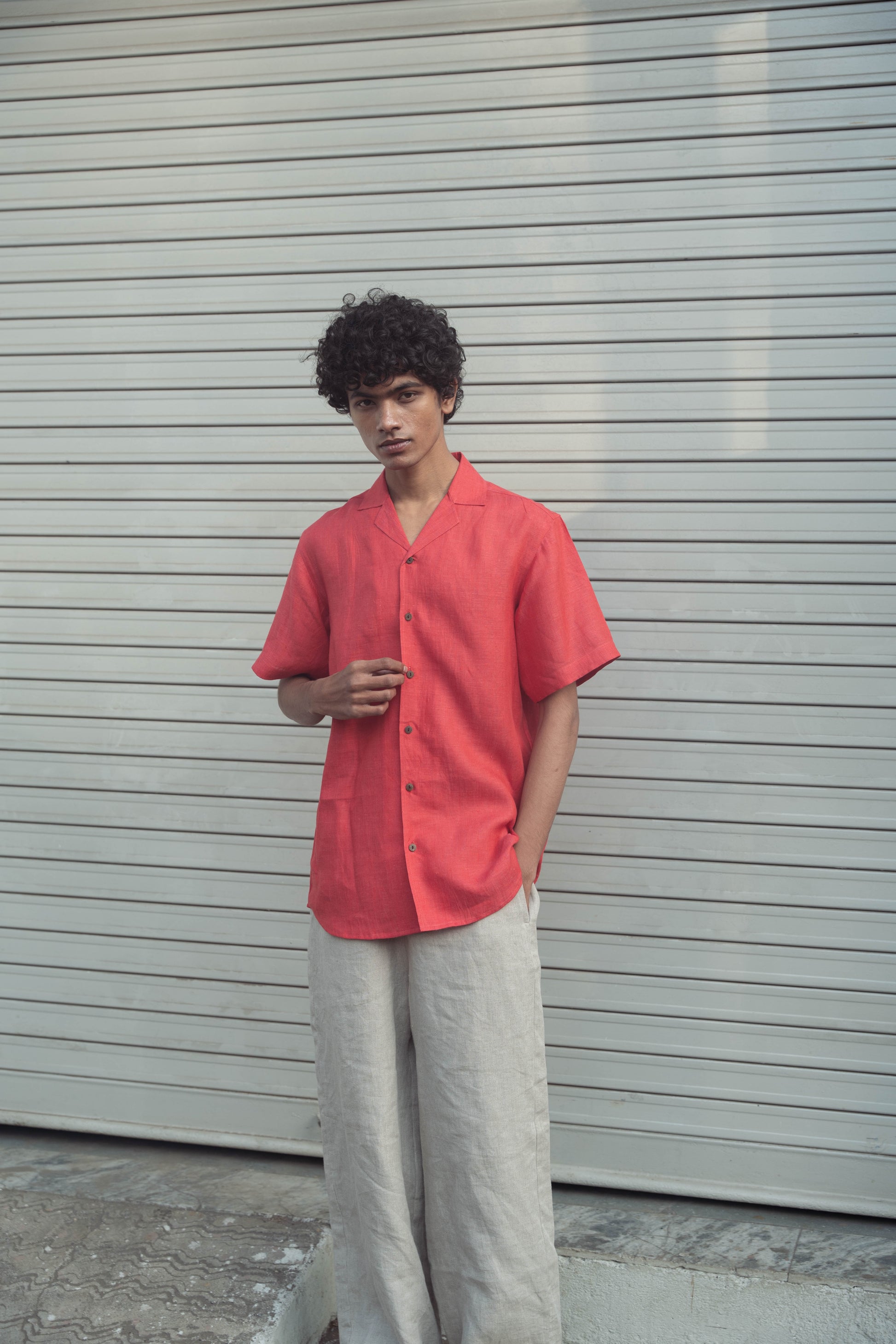 A man in a rich red linen half-sleeved shirt and beige pants is standing in front of a white shutter shop