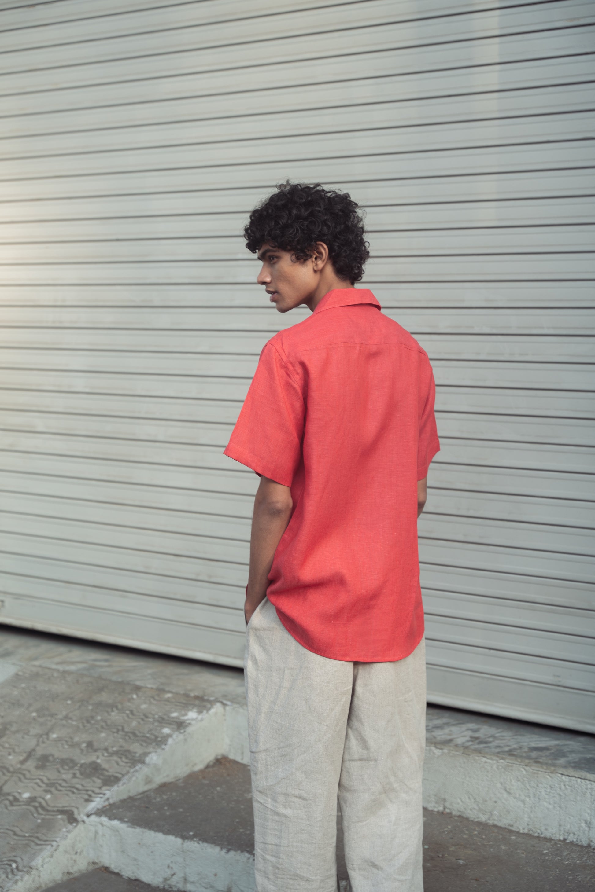 Back shot of a man in a rich red linen half-sleeved shirt and beige pants is standing in front of a white shutter shop