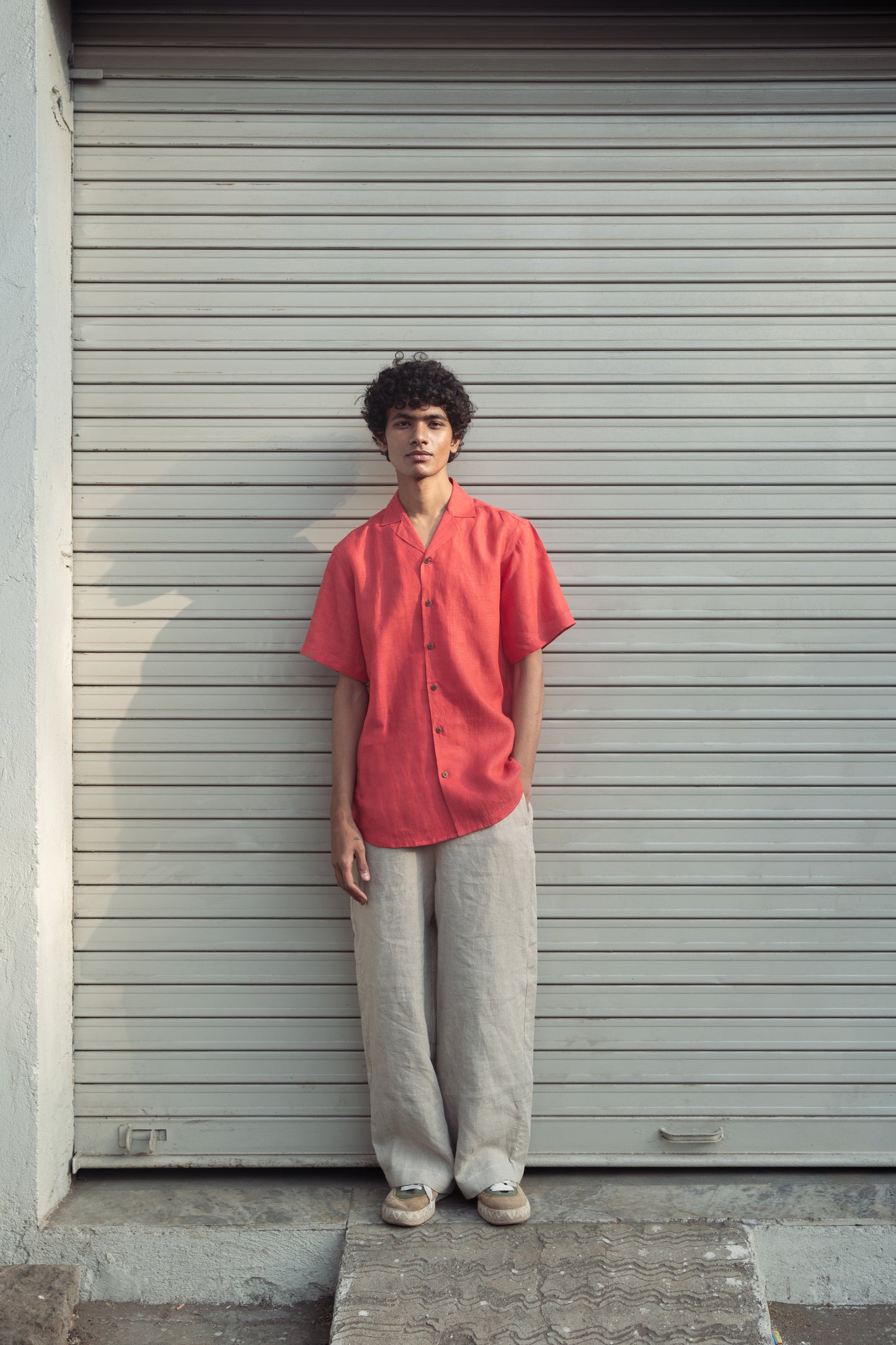 A man in a rich red linen half-sleeved shirt and beige pants is standing in front of a white shutter shop