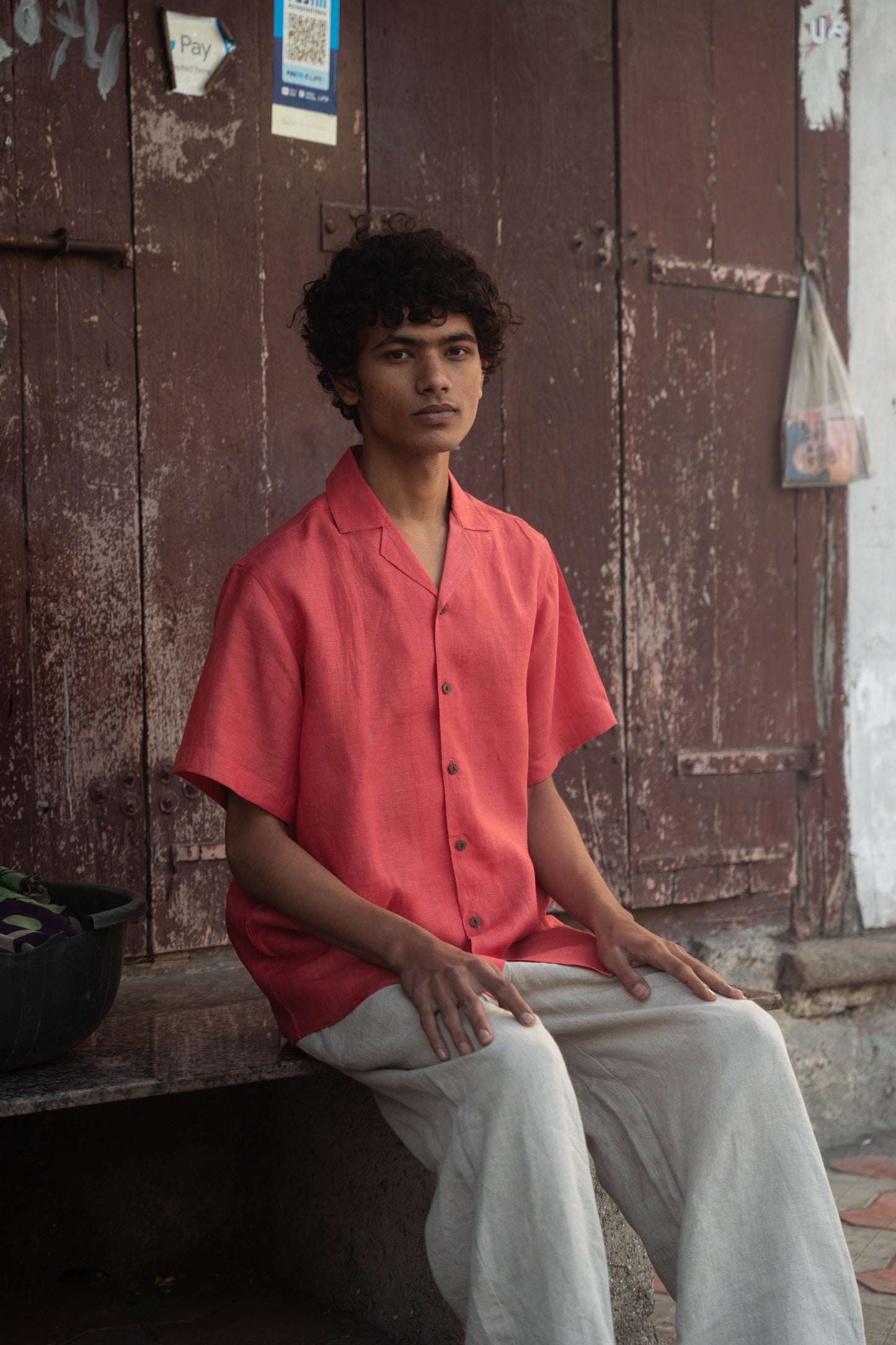 closer shot of a man in a rich red linen half-sleeved shirt is sitting in front of a wide brown door