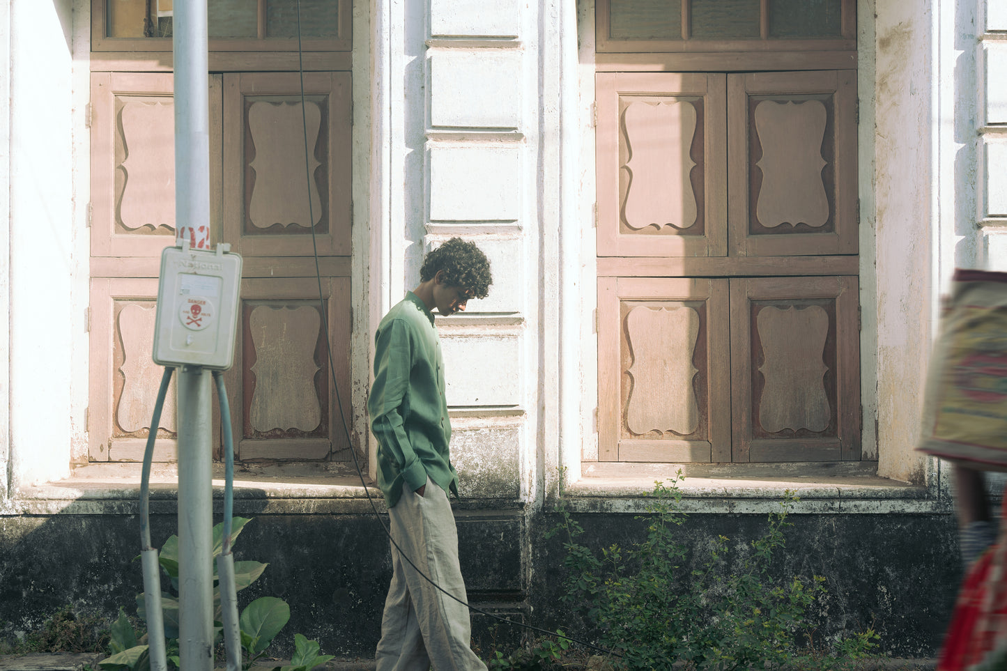 A man in a green pure linen full-sleeved shirt and beige linen pants walking infront of an old house