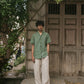 A man in a green pure linen half-sleeved shirt and beige linen pants standing infront of an old door