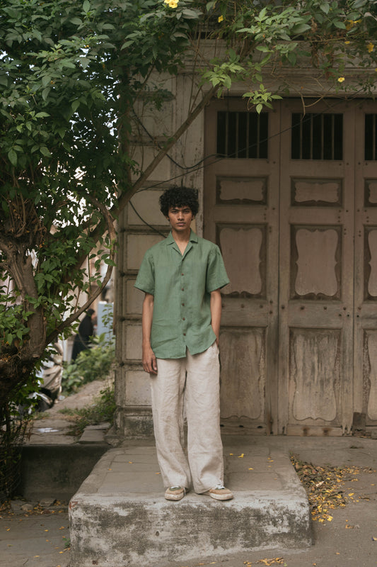 A man in a green pure linen half-sleeved shirt and beige linen pants standing infront of an old door