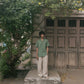 A wider shot of a man in a green pure linen half-sleeved shirt and beige linen pants standing infront of an old house