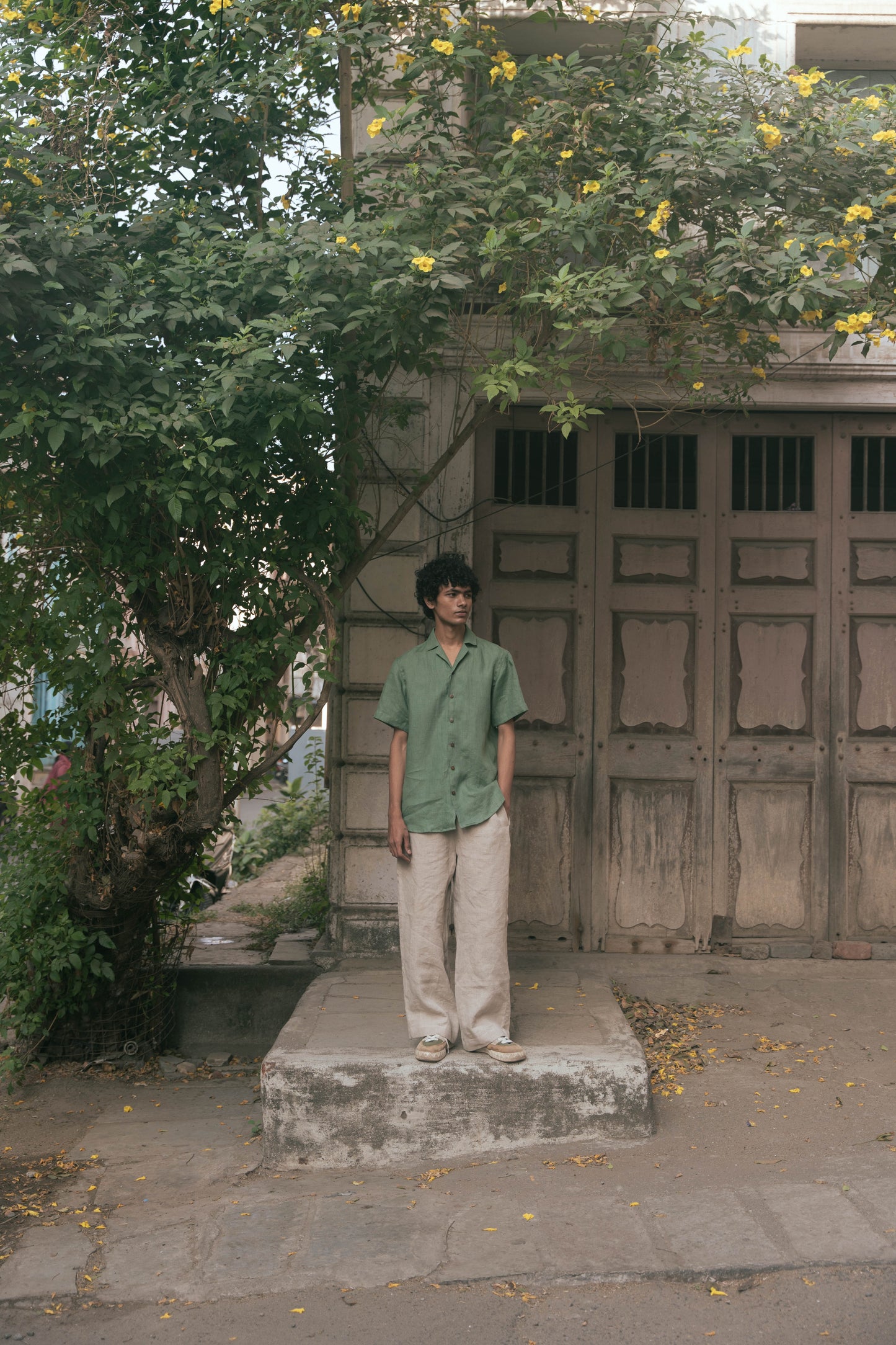 A wider shot of a man in a green pure linen half-sleeved shirt and beige linen pants standing infront of an old house