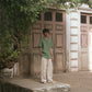 A wider shot of a man in a green pure linen half-sleeved shirt and beige linen pants standing infront of an old house