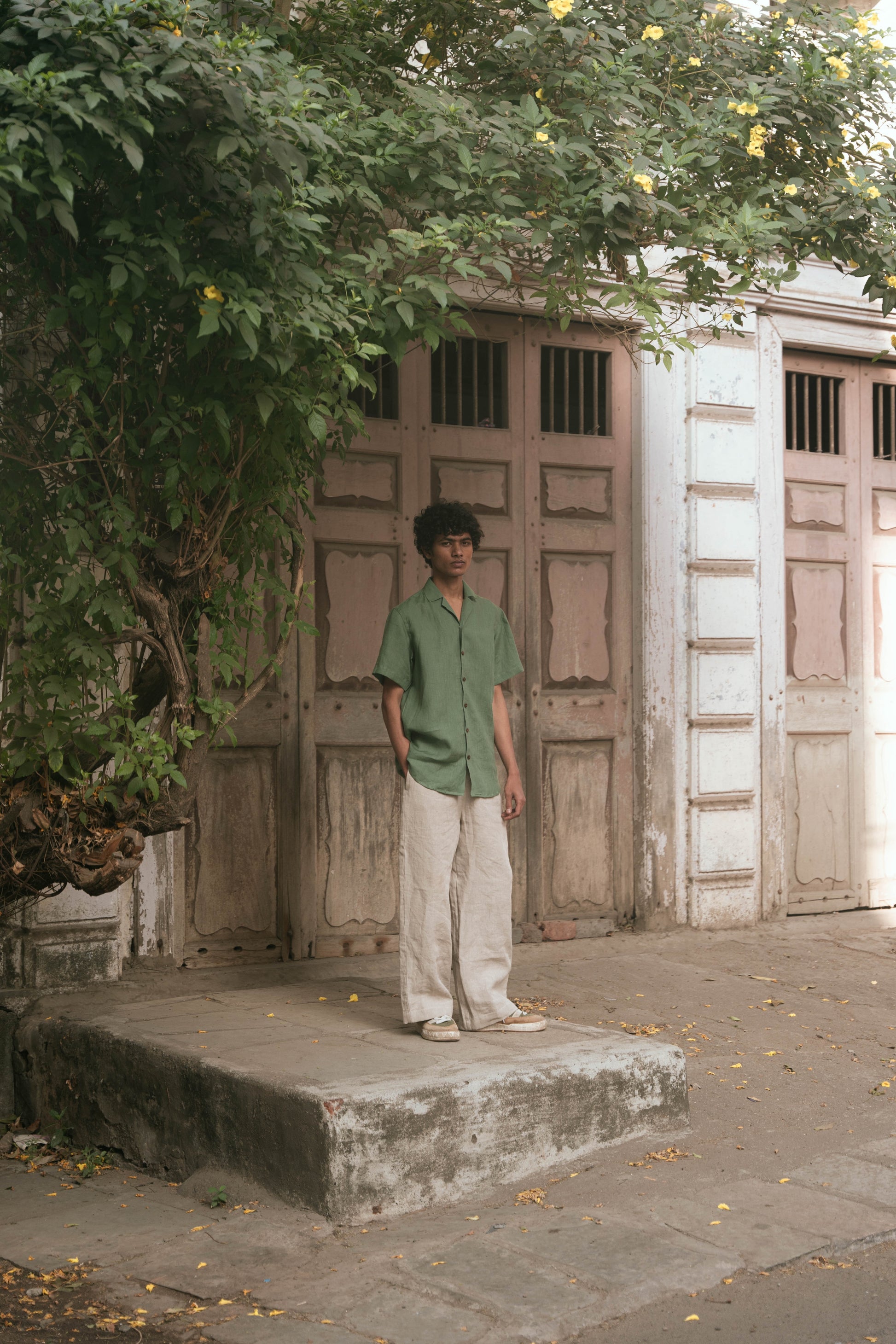 A wider shot of a man in a green pure linen half-sleeved shirt and beige linen pants standing infront of an old house