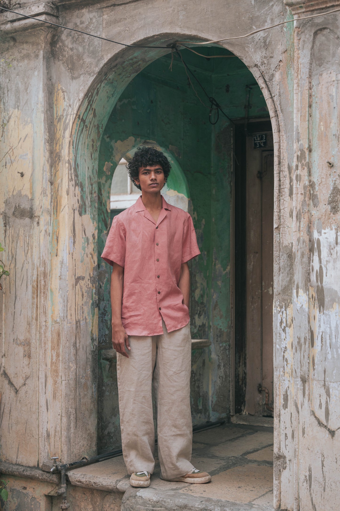 A man in peach linen half-sleeved cuban collared shirt standing in front of a door to a rustic house