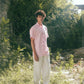 A man in light pink half-sleeved cuban collared linen shirt and white linen pants in between green grass and bushes