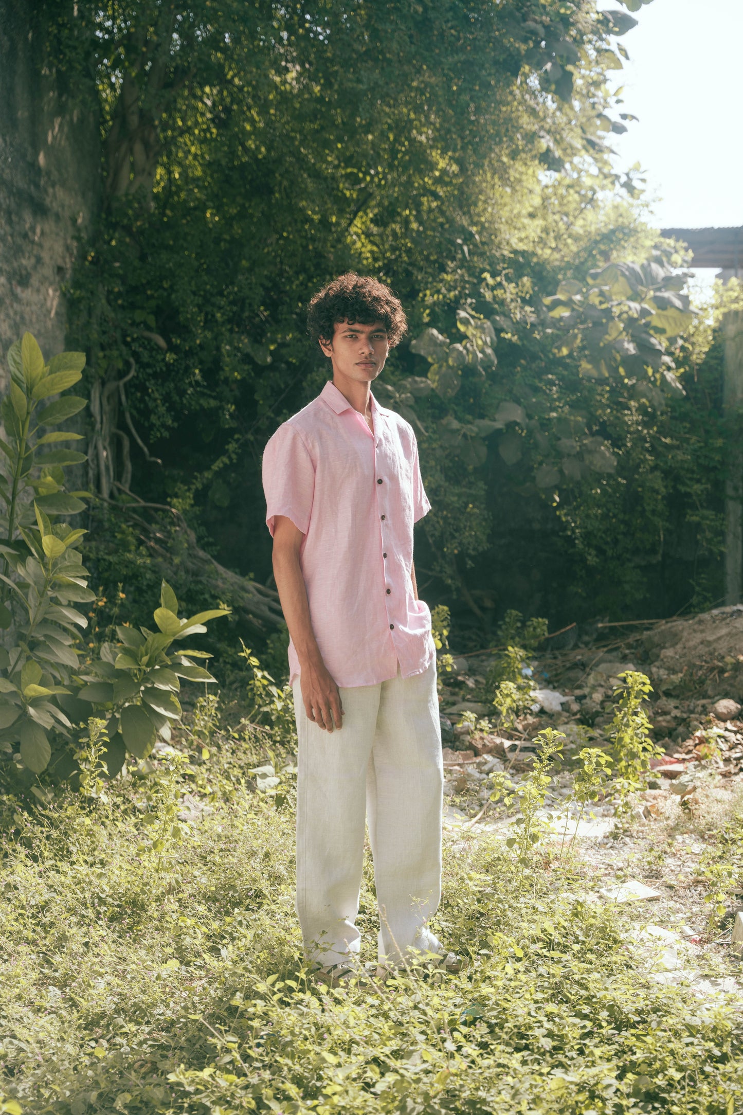 A man in light pink half-sleeved cuban collared linen shirt and white linen pants in between green grass and bushes