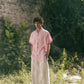 A man in light pink half-sleeved cuban collared linen shirt and white linen pants in between green grass and bushes