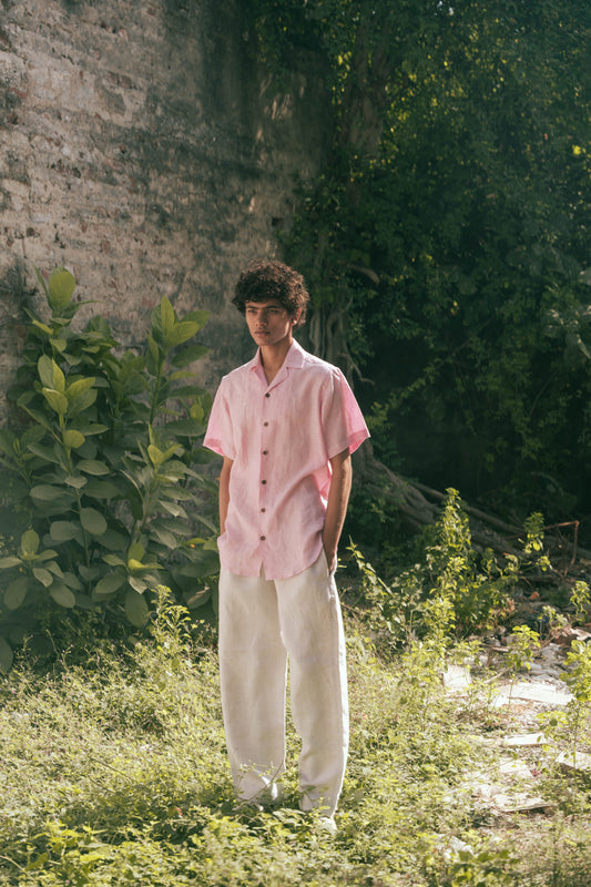 A man in light pink half-sleeved cuban collared linen shirt and white linen pants in between green grass and bushes