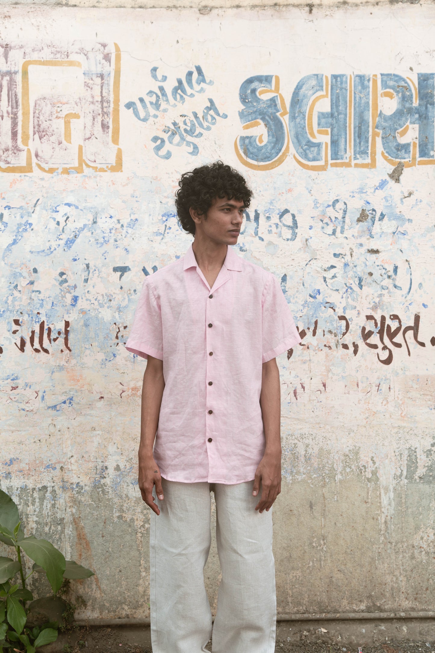 A man in light pink half-sleeved cuban collared linen shirt and white linen pants in front of a graffiti wall