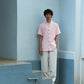 A man in light pink half-sleeved cuban collared linen shirt and white linen pants standing in front of a blue wall