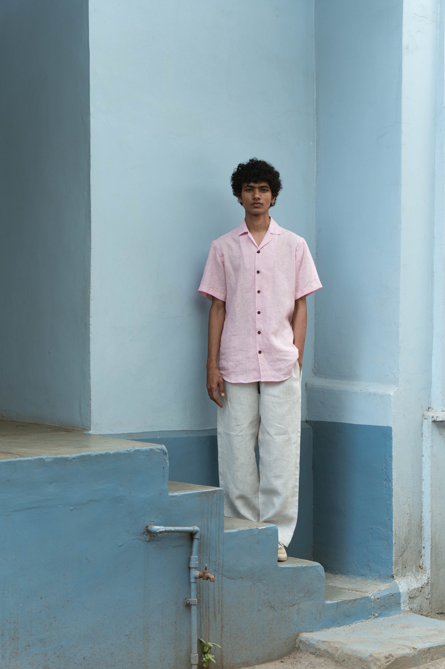 A man in light pink half-sleeved cuban collared linen shirt and white linen pants standing in front of a blue wall