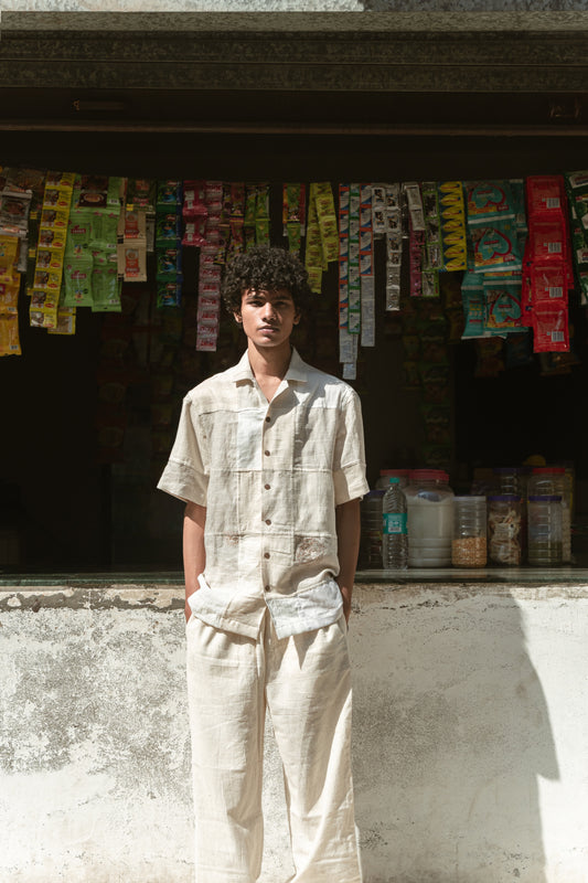 A young man in an off-white half-sleeved shirt made with patches of cotton and linen squares is standing in front of a shop