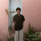 A man in a black linen half-sleeve regular collar shirt is standing infront a pink wall with a blue door