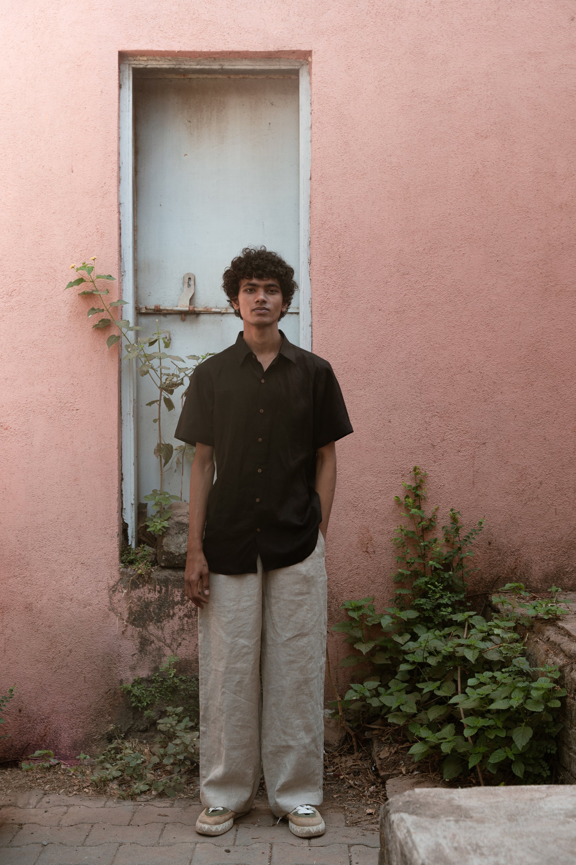 A man in a black linen half-sleeve regular collar shirt is standing infront a pink wall with a blue door