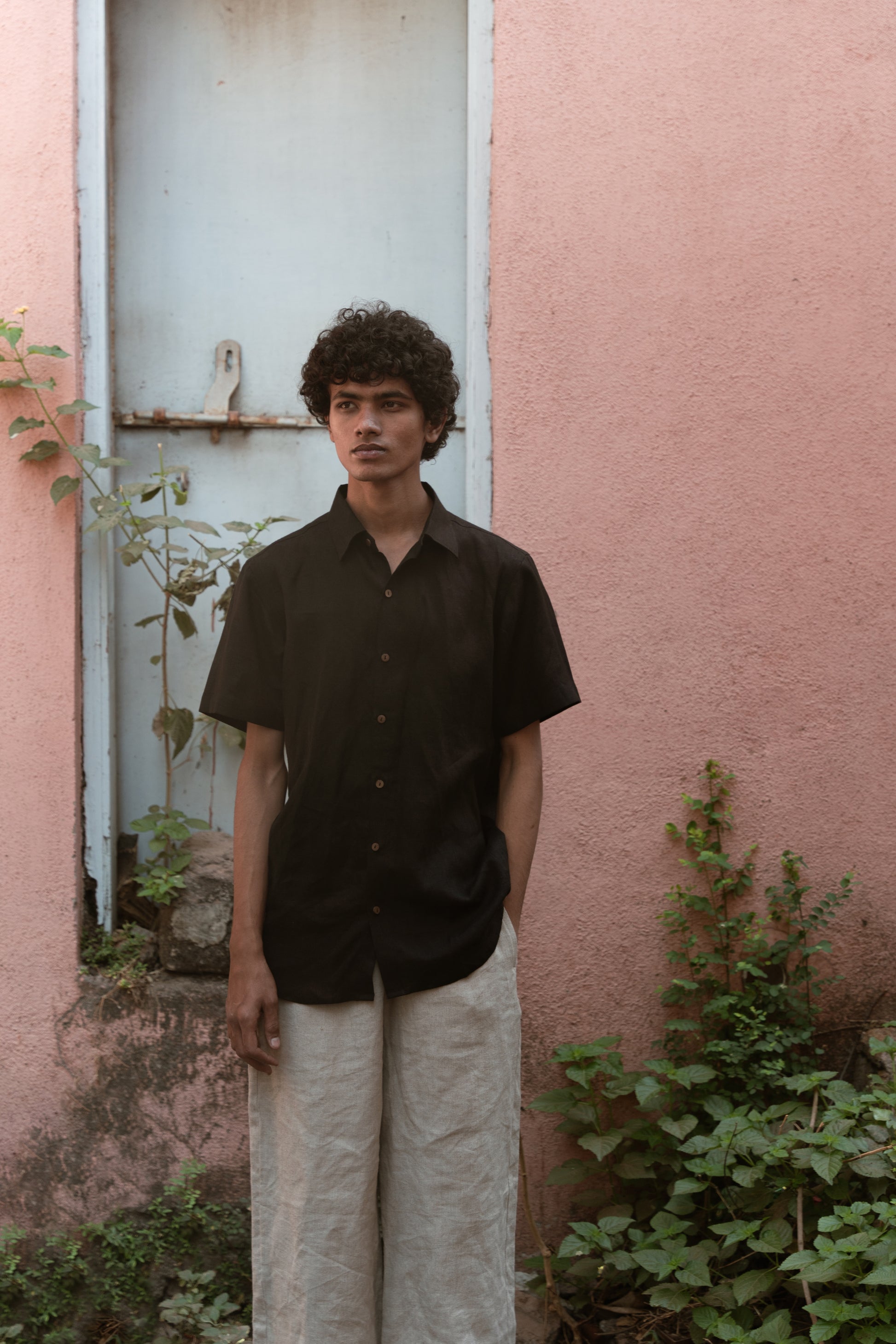 Closer look of a man in a black linen half-sleeve regular collar shirt is standing infront a pink wall with a blue door