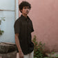 A man in a black linen half-sleeve regular collar shirt is standing infront a pink wall looking straight in the camera