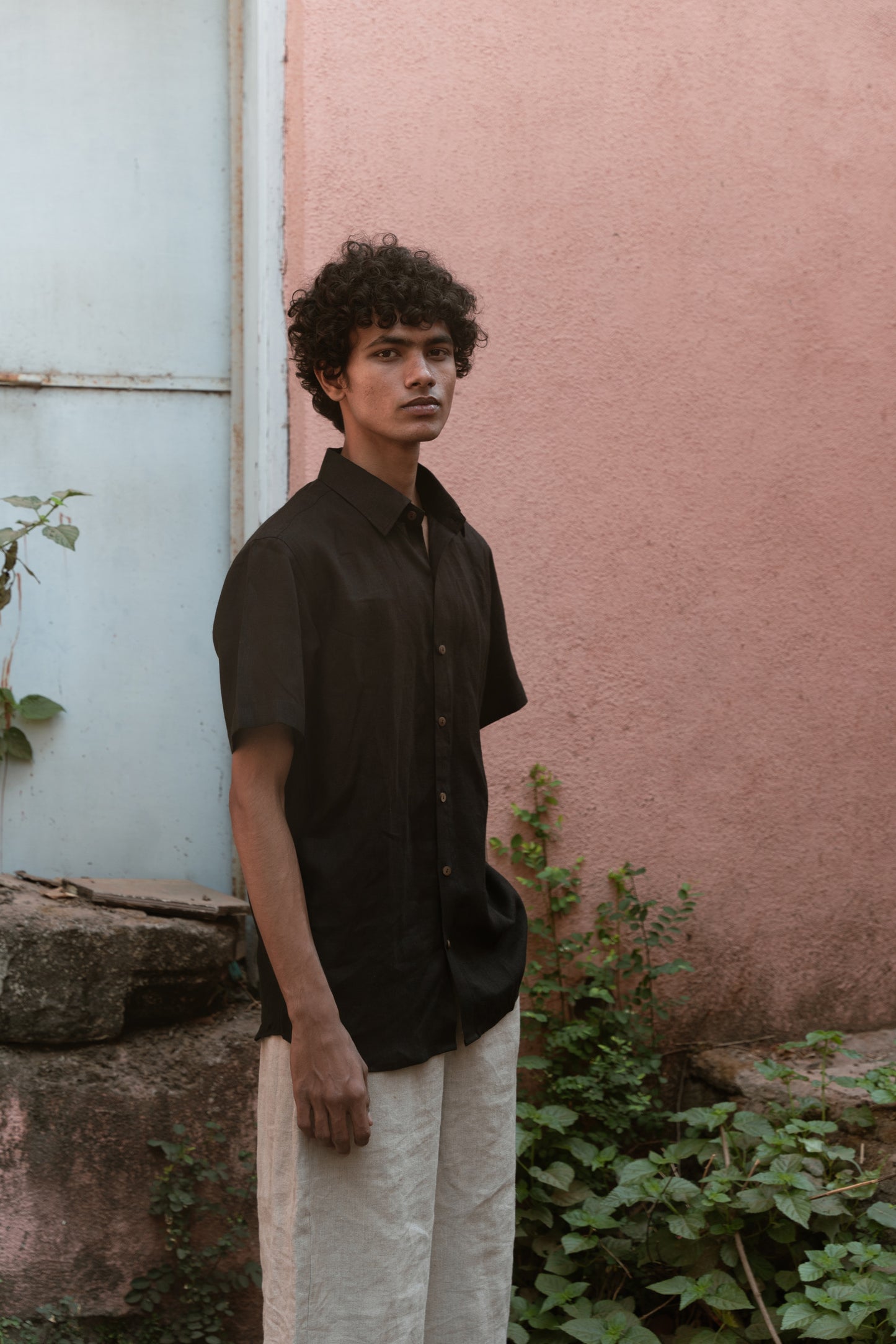 A man in a black linen half-sleeve regular collar shirt is standing infront a pink wall looking straight in the camera