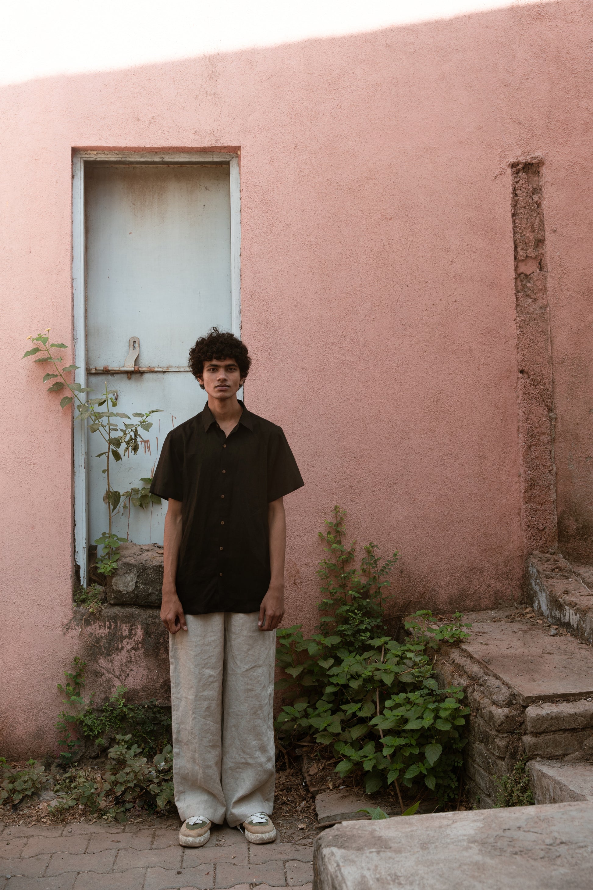 A man in a black linen half-sleeve regular collar shirt and beige linen pants is standing infront a pink wall with a blue door