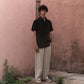 A man in a black linen half-sleeve regular collar shirt is standing infront a pink wall 