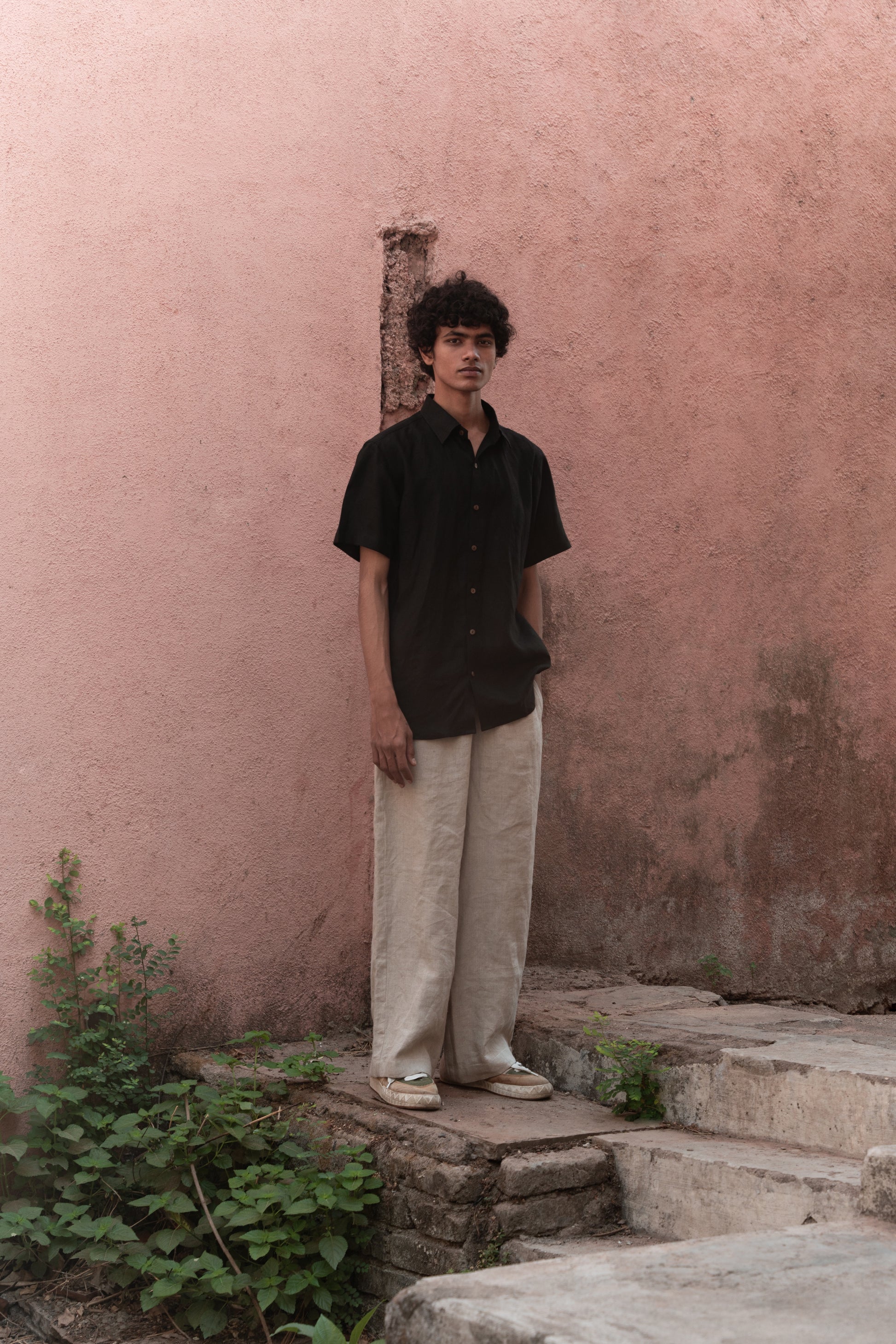 A man in a black linen half-sleeve regular collar shirt is standing infront a pink wall 