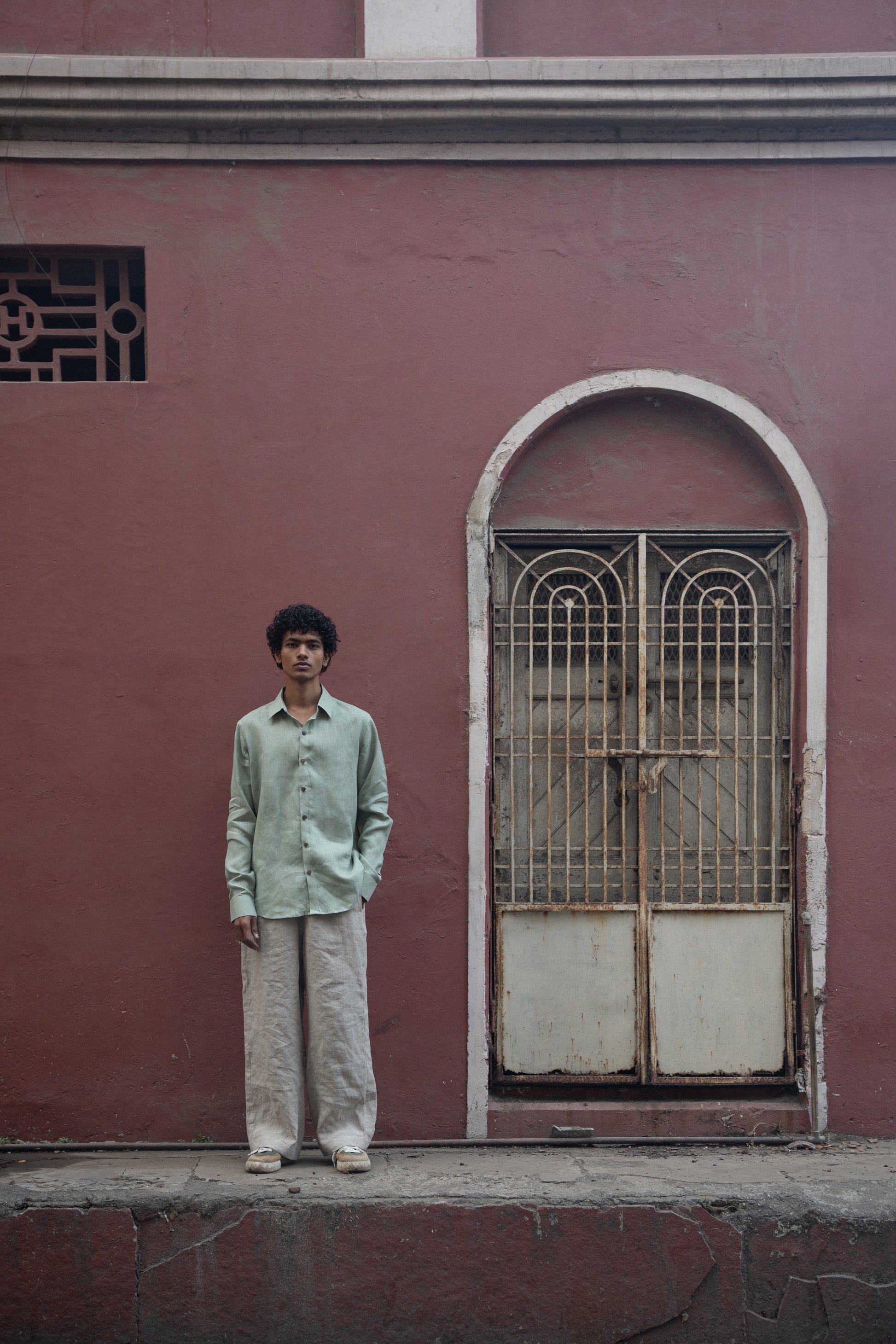 Wider shot of A man in mint-green pure linen full-sleeved shirt and light beige linen pants is standing in front of a pink wall