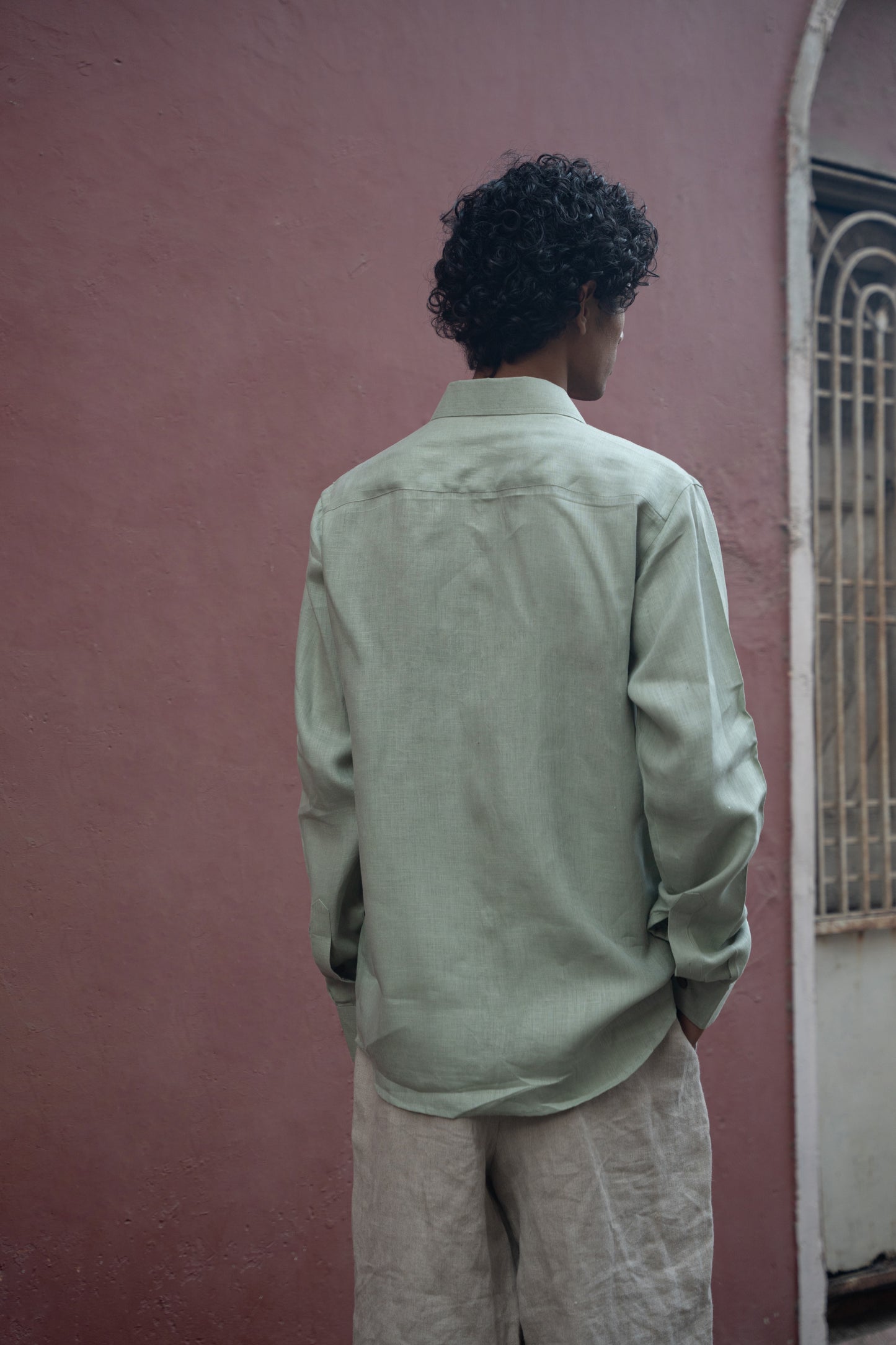 Back shot of man in mint-green pure linen full-sleeved shirt is standing in front of a pink wall