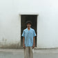 A man in blue linen cuban collared half sleeve shirt with off-white pants is standing in front of a white wall