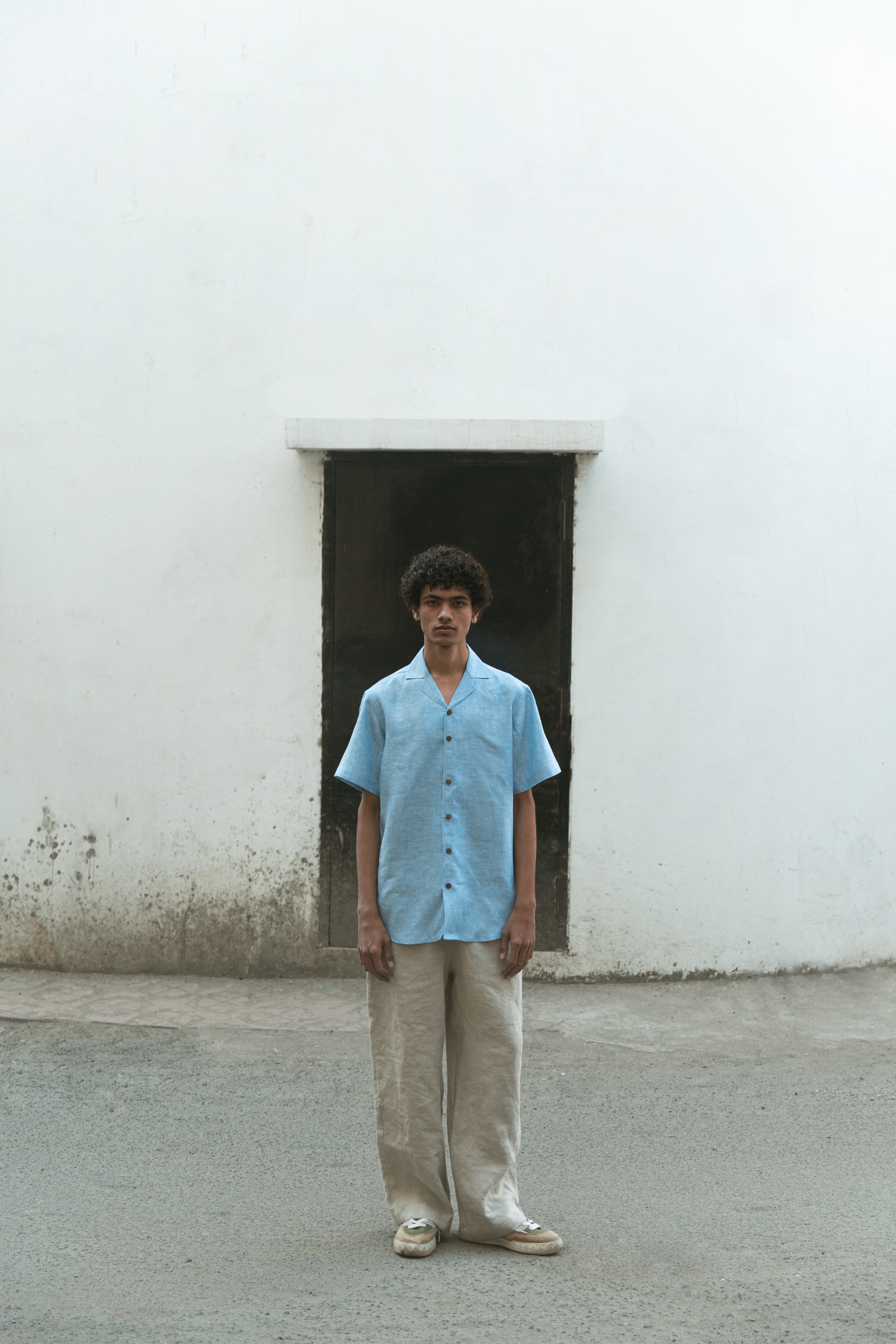 A man in blue linen cuban collared half sleeve shirt with off-white pants is standing in front of a white wall