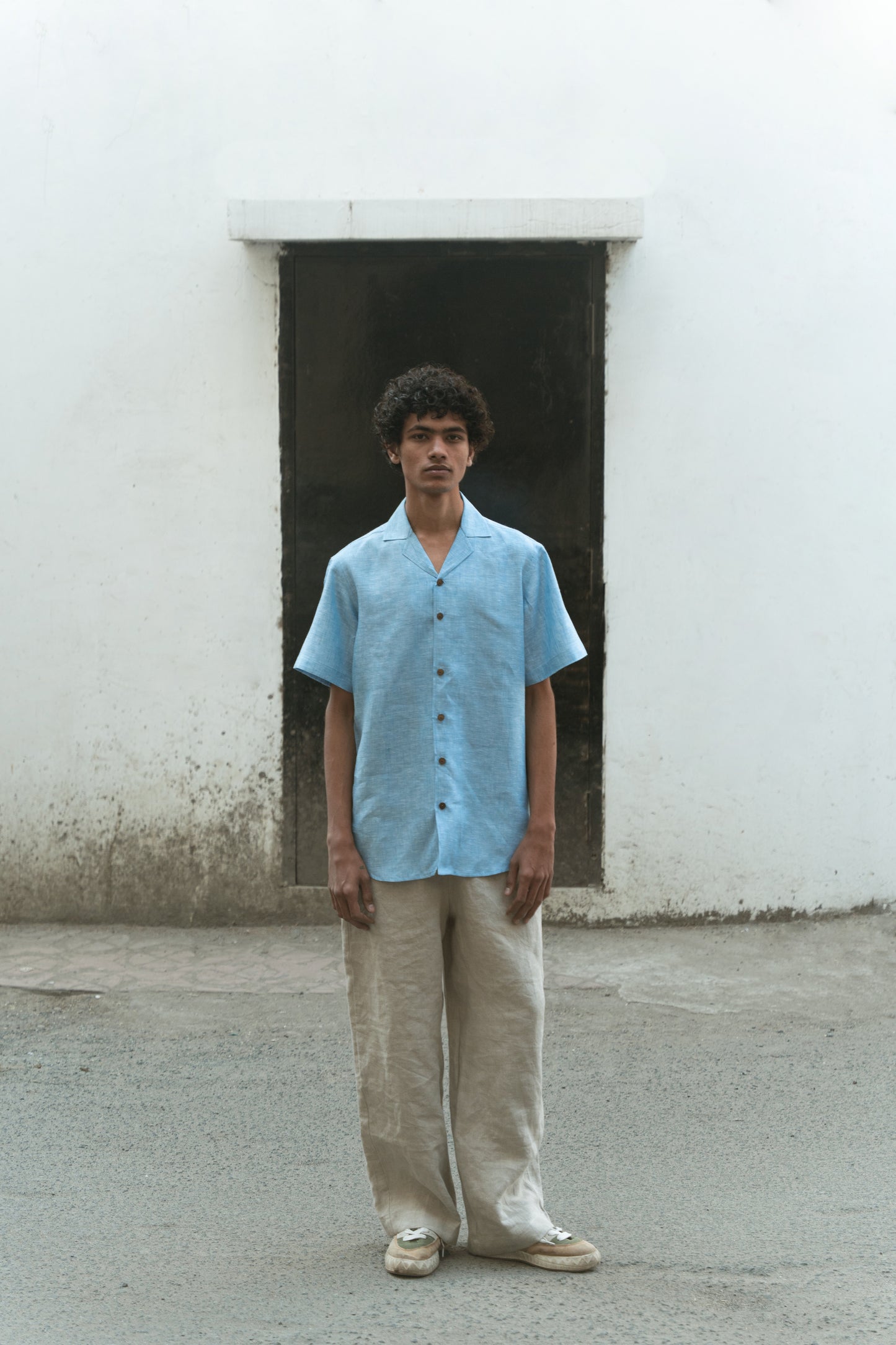 A man in blue linen cuban collared half sleeve shirt is standing in front of a off white wall