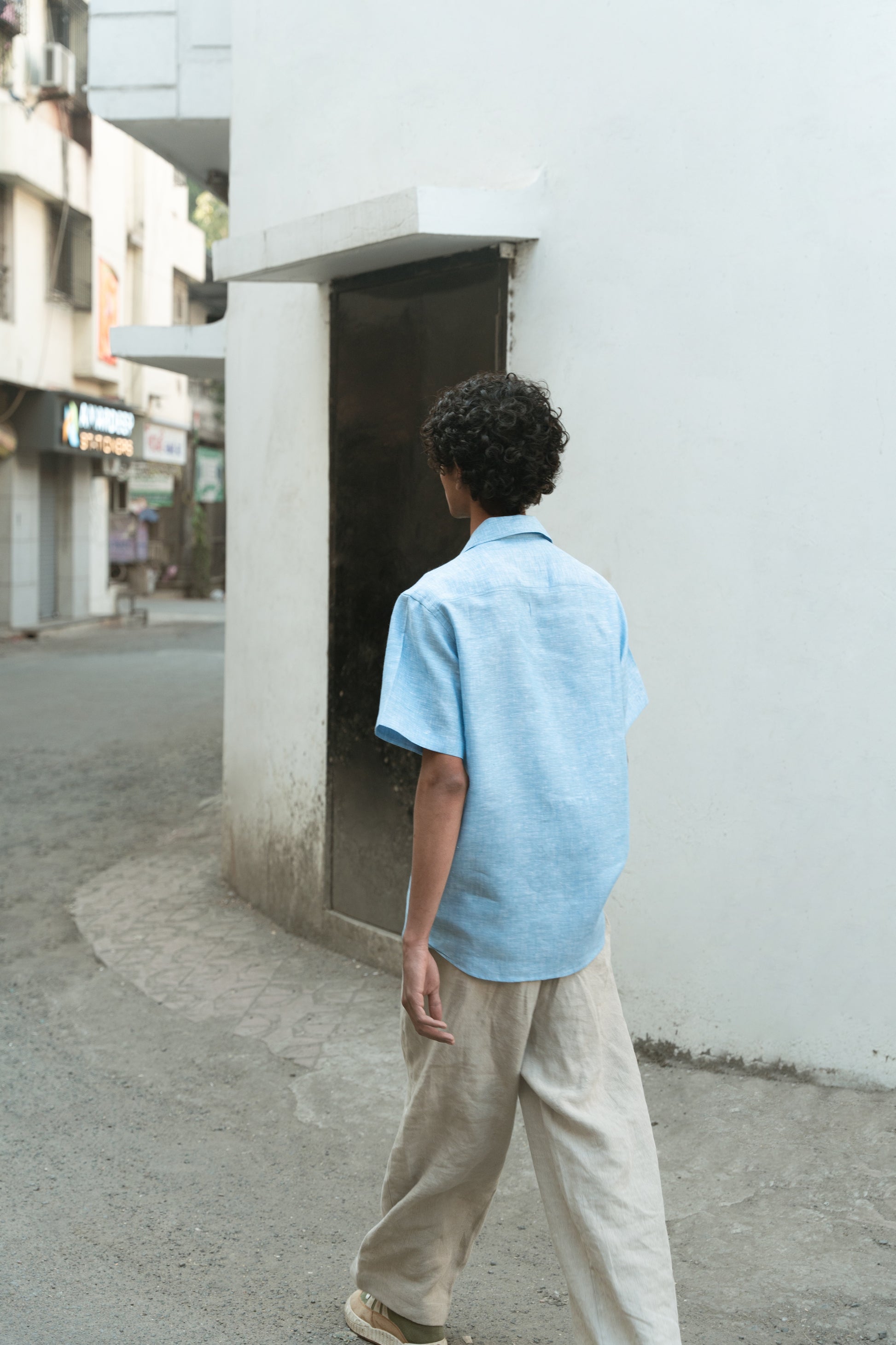 Back shot of a man in a blue linen shirts and off-white pants walking on the streets