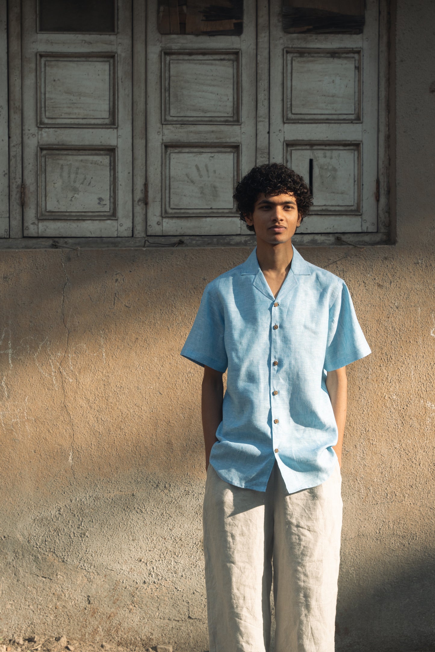 A man in blue linen cuban collared half sleeve shirt is standing in front of a light beige wall