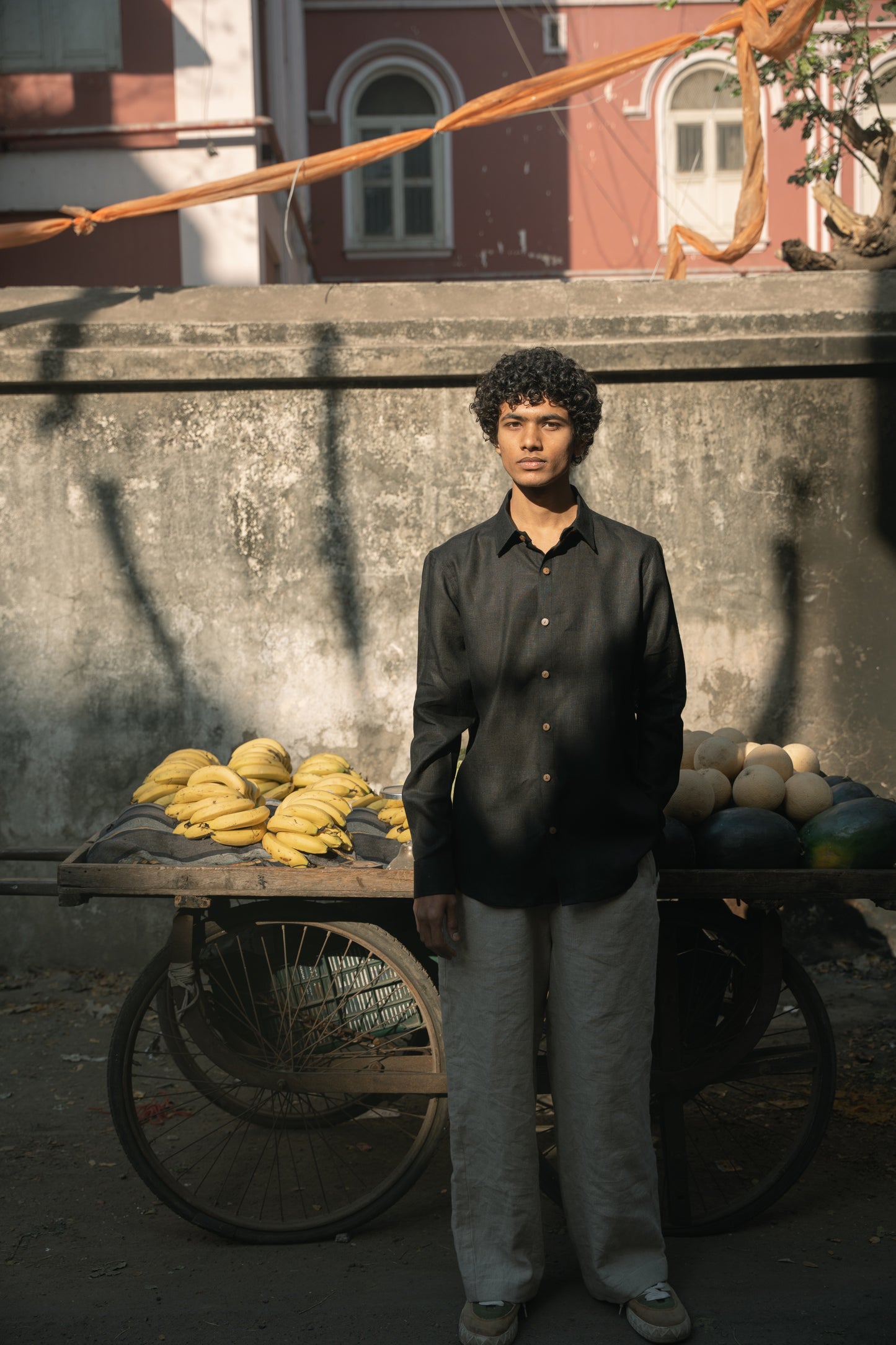 A man in a black linen full-sleeve regular collar shirt is standing in front of a cart with bananas and melons