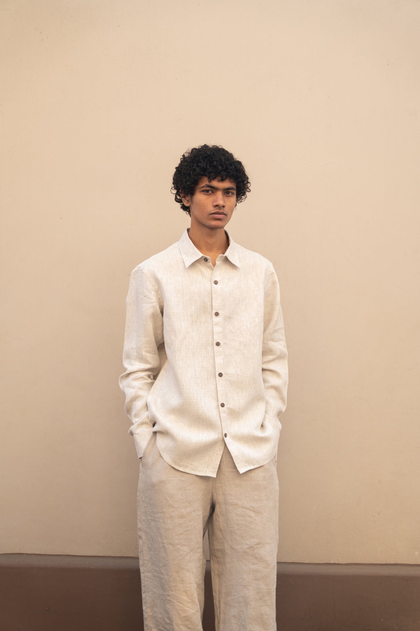 A man in light beige full sleeved pure linen shirt and a darker beige pants is standing in front  of a wall