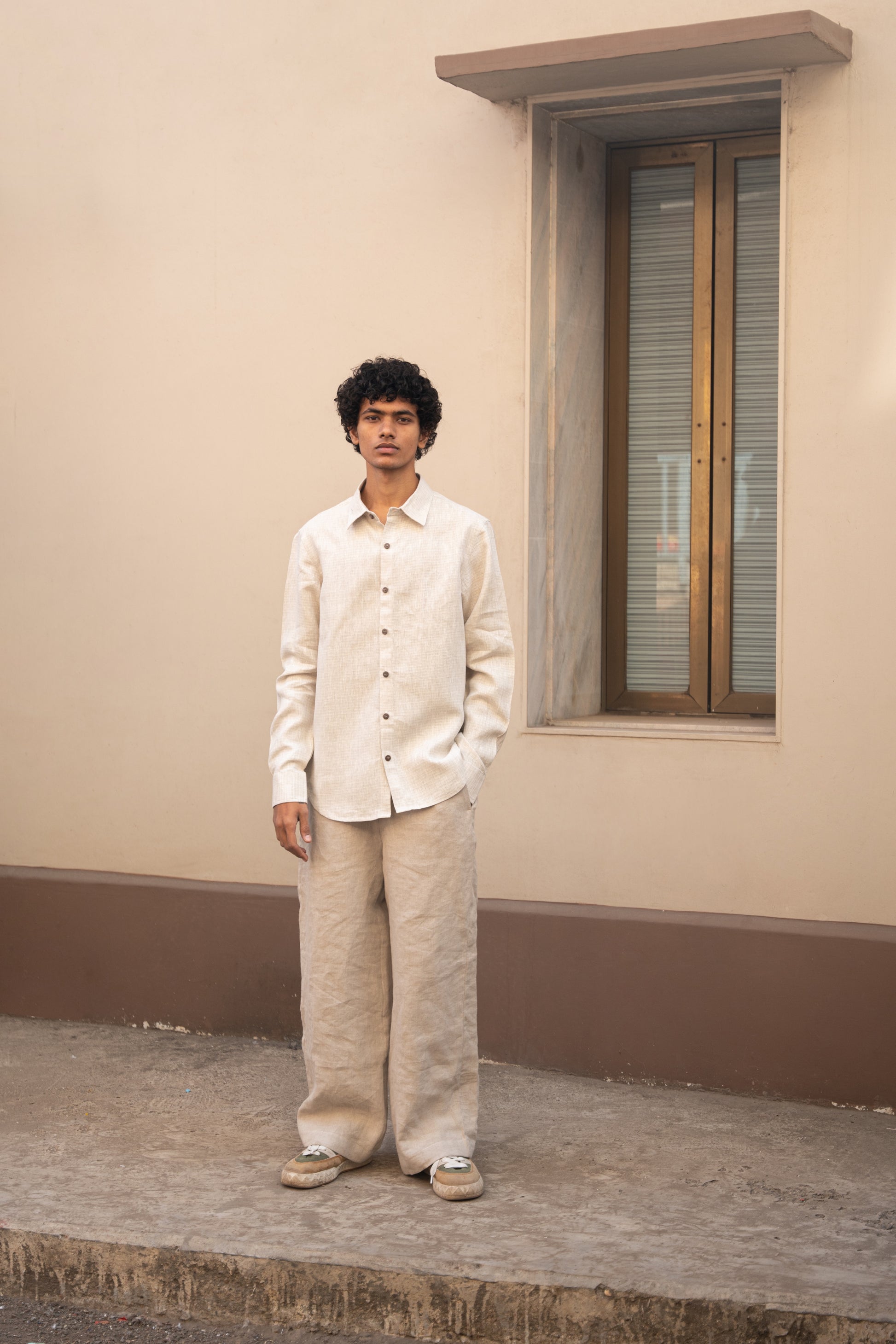 A wider shot of a man in light beige full sleeved pure linen shirt and a darker beige pants standing in front  of a wall