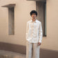 A man in light beige full sleeved pure linen shirt and a darker beige pants is standing in front  of a wall